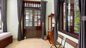 A room with a bed, a wooden chair, a small table, a window with curtains, and a door with a view of trees. - Vi TamCoc Hotel (Nam Hoa Lu)