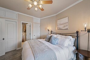 Un dormitorio con cama, ventilador de techo, mesita de noche, lámpara de pared y una puerta que da a otra habitación.