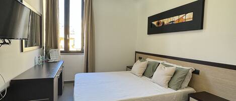 A modern hotel room with a bed, bedside tables, a desk, a TV, and a window with curtains.