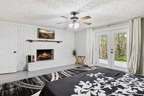 A bedroom with a fireplace, a bed with a black and white patterned bedspread, a ceiling fan, and a view of the outdoors through large windows.