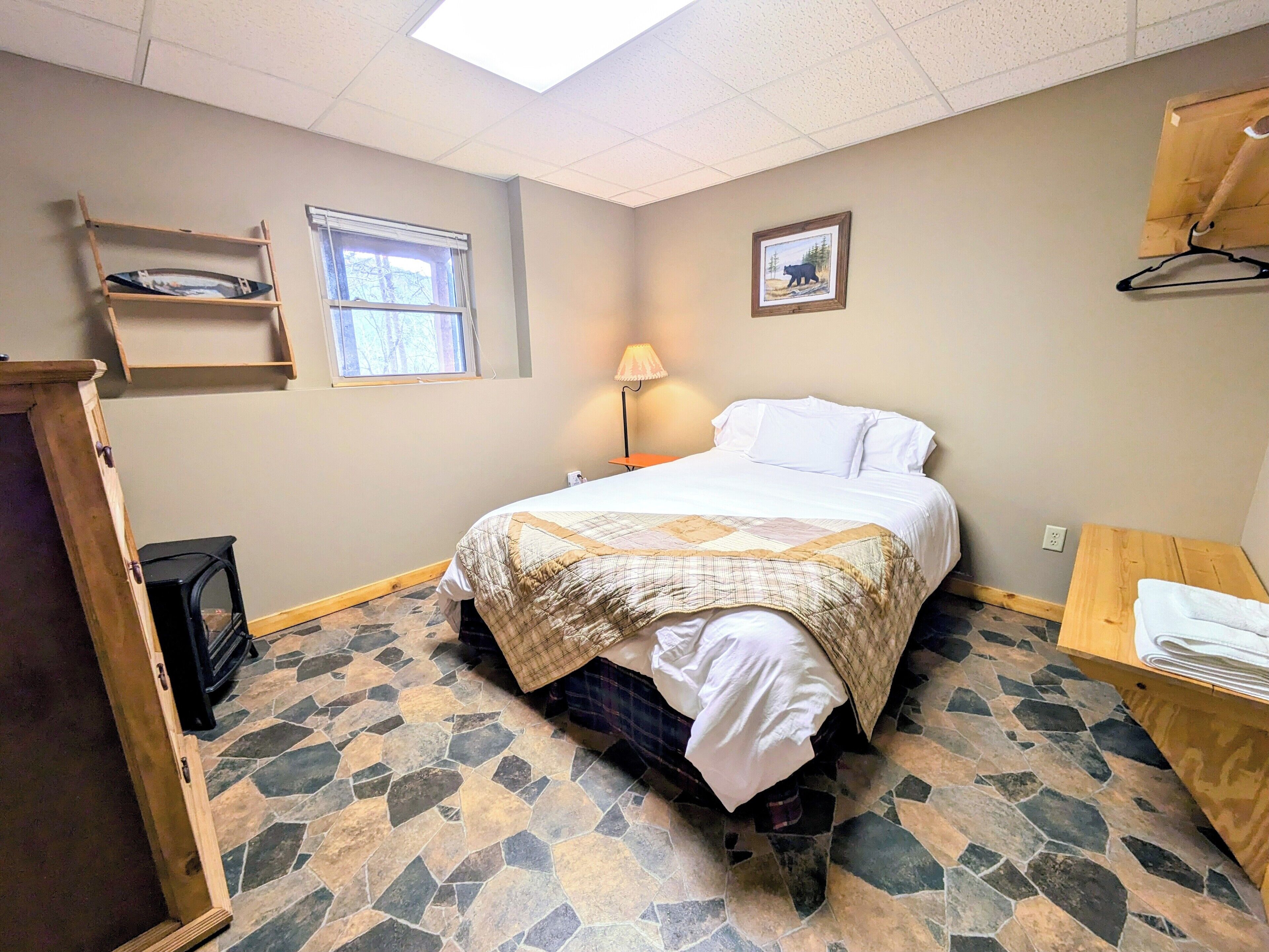 Un dormitorio con cama, ventana, lámpara, estante de madera y un cuadro en la pared.