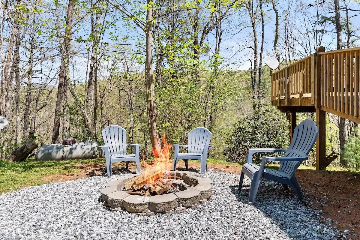 A fire pit with logs, surrounded by four Adirondack chairs, set on gravel.