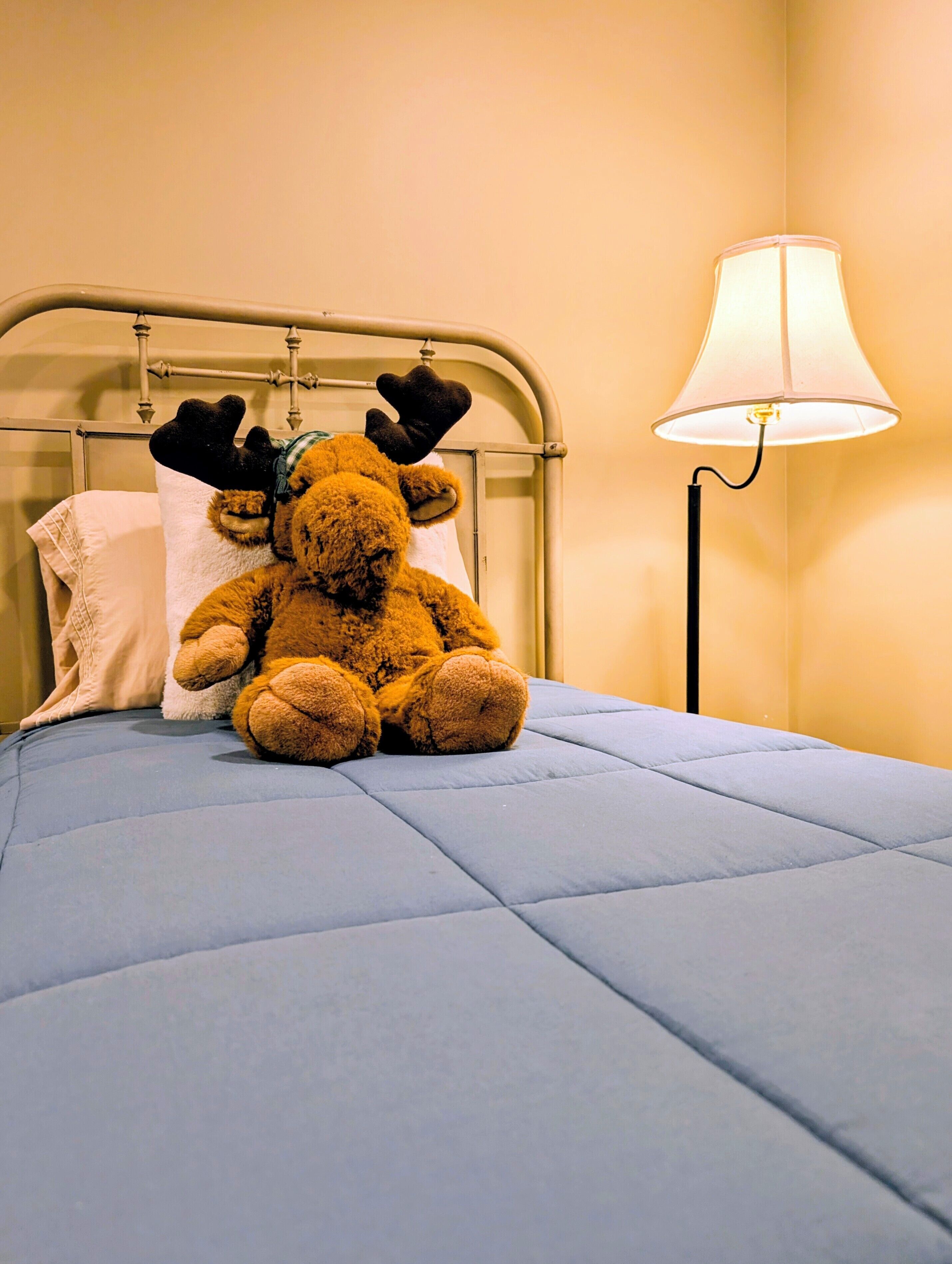 Uma cama com colchão azul, um urso de pelúcia, travesseiro branco e uma luminária.