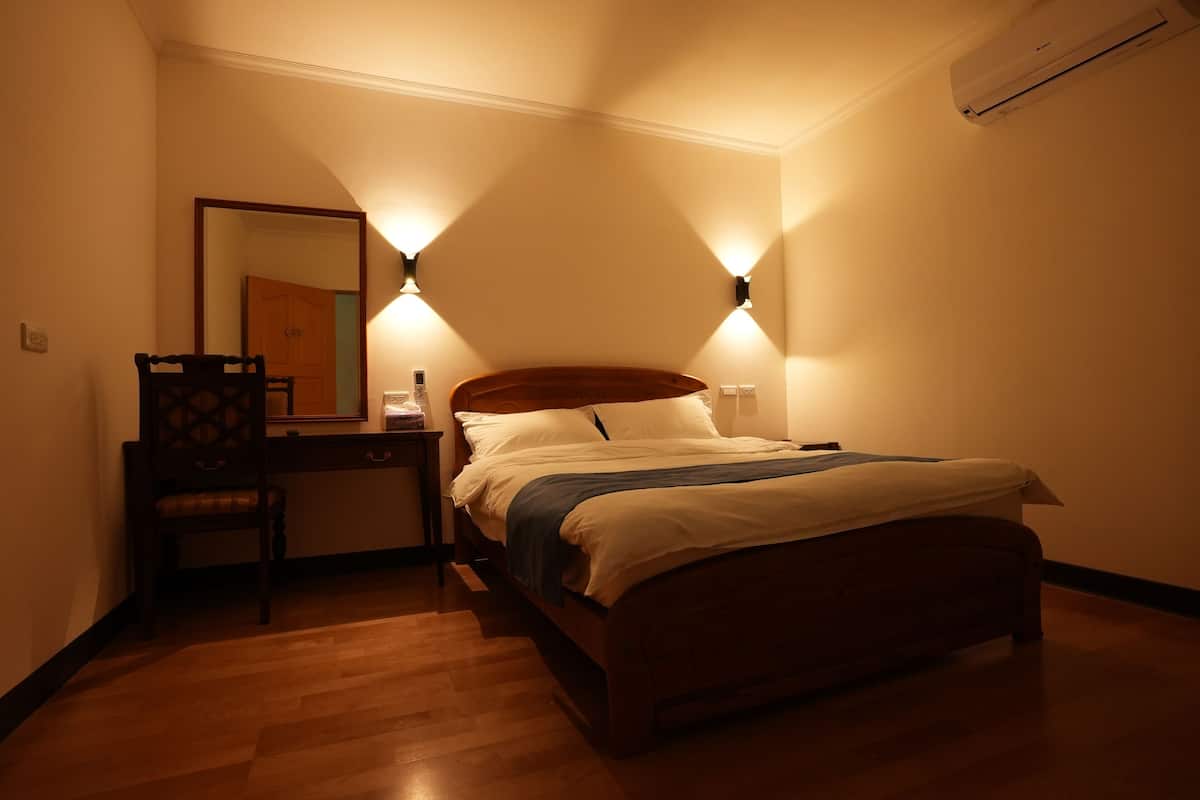 Quarto com cama de madeira, mesa de cabeceira, cadeira e duas luzes fixadas na parede.