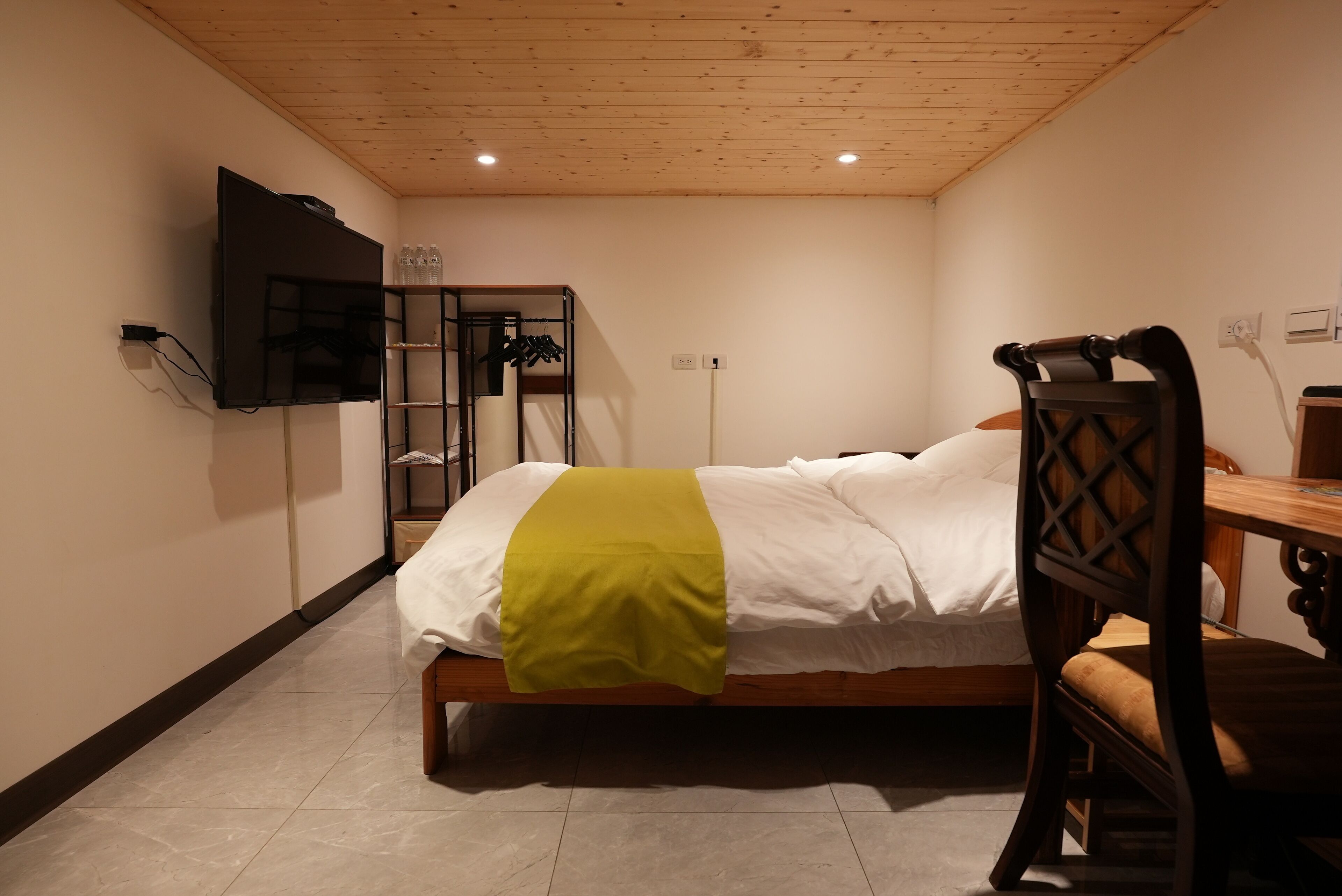 A bedroom with a bed, a wooden chair, a desk, a television mounted on the wall, and a shelf with bottles.