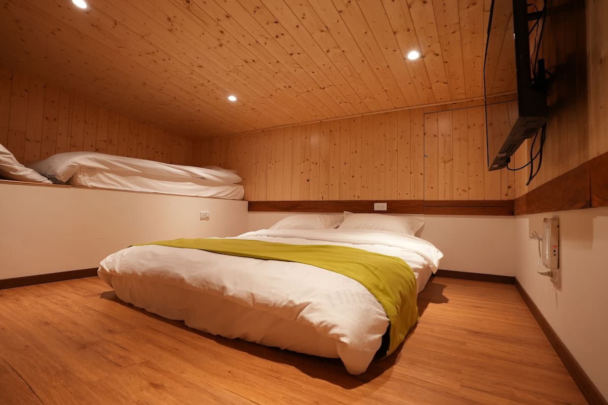 Quarto com piso de madeira, cama com roupa de cama branca e manta verde, e teto de madeira com iluminação embutida.