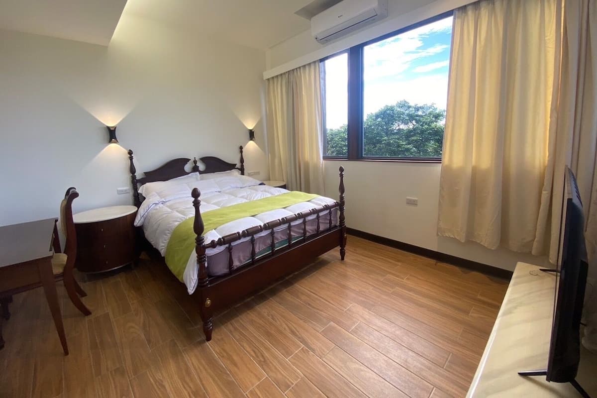 Quarto com cama de madeira, duas luminárias de cabeceira, janela com cortinas e ar condicionado.