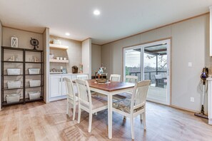 A dining area with a wooden table and white chairs, a kitchen with white cabinets and shelves, and a sliding glass door leading to an outdoor deck.