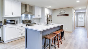 A modern kitchen with white cabinets, a stainless steel range hood, a microwave, and a large island with bar stools. - Hudson Oaks Country Creekside Home: Hot Tub & Deck (Weatherford)