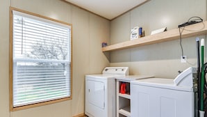 A laundry room with a washer and dryer, wooden shelves, and a window with blinds. - Hudson Oaks Country Creekside Home: Hot Tub & Deck (Weatherford)