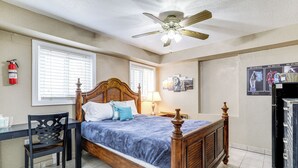 Un dormitorio con una cama de madera, un escritorio, una silla, un ventilador de techo y un extintor.