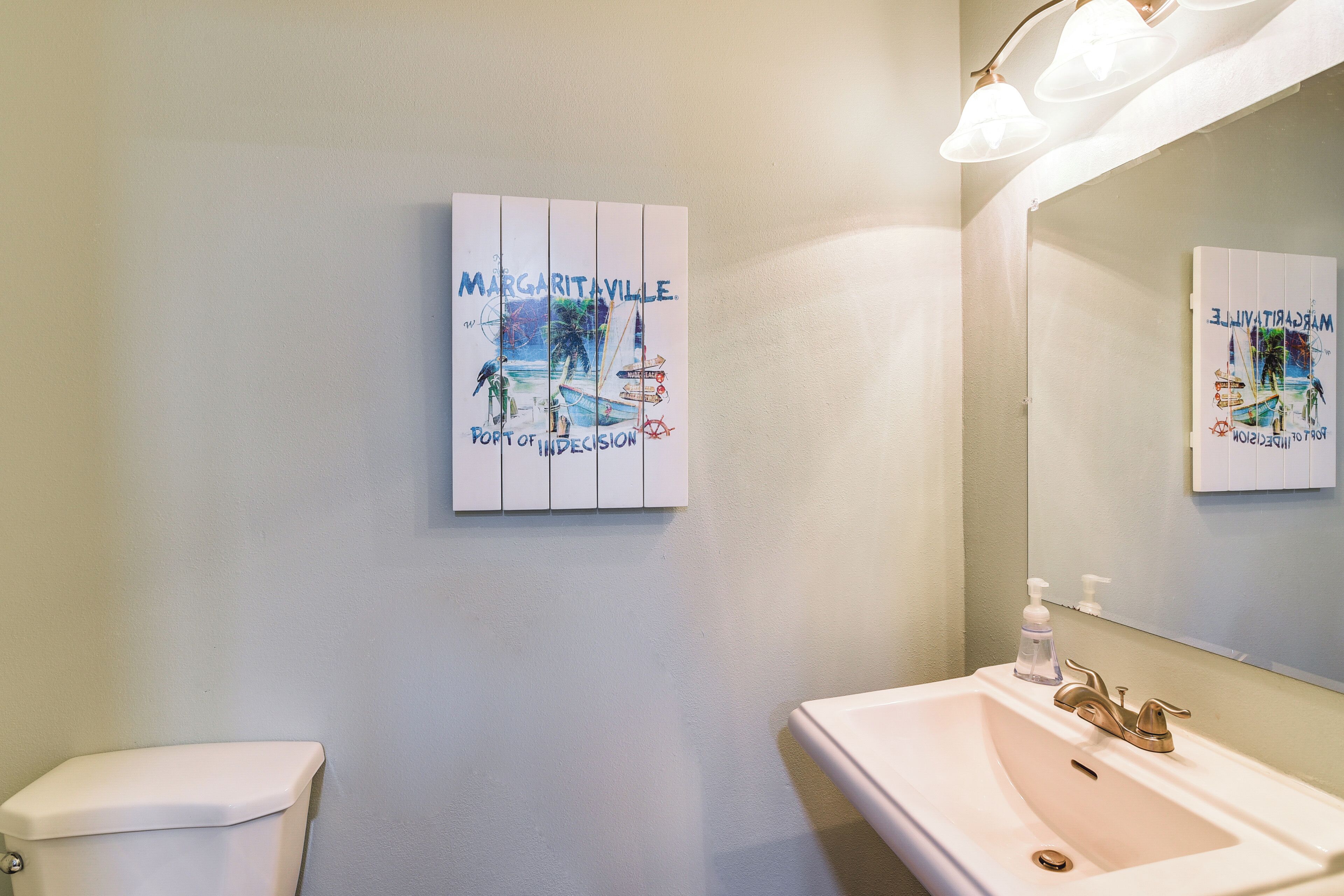 Et badeværelse med en hvid vask, toilet og et spejl monteret på væggen. Der er et billede i ramme med et strandmotiv samt teksten "Margaritaville Port of Decision".