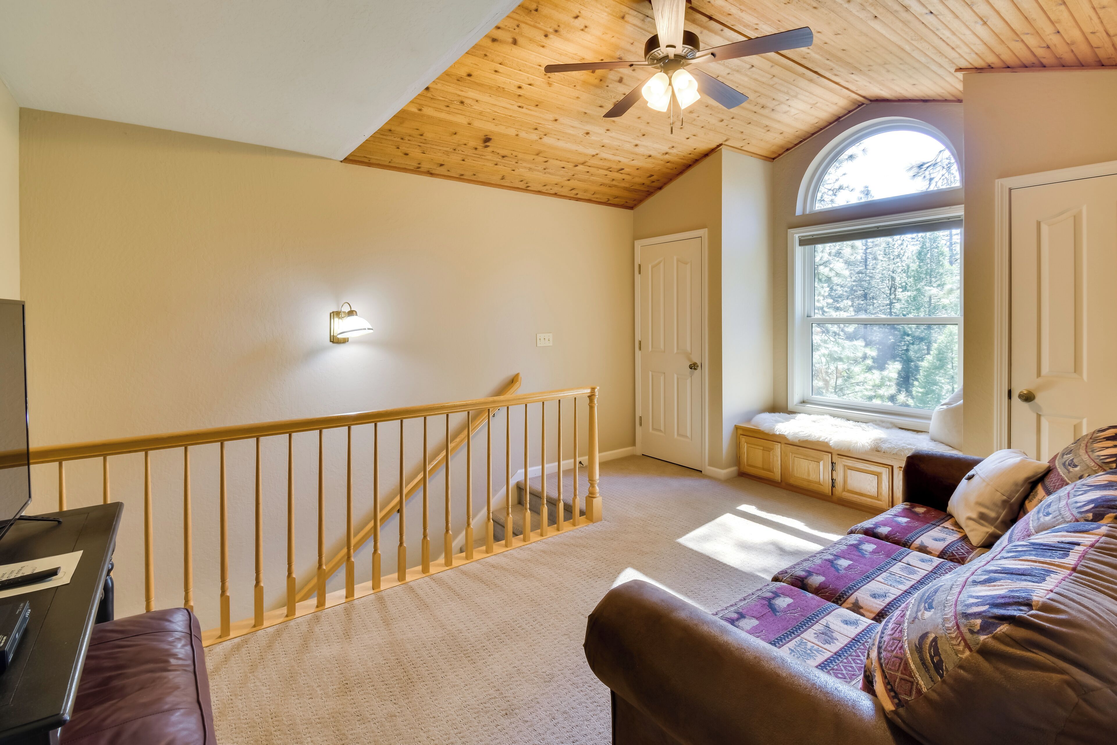 Ein gemütliches Zimmer mit Holzbalkendecke, einer Treppe, einer Bank neben einem großen Fenster und einem Sofa mit gemusterten Kissen.