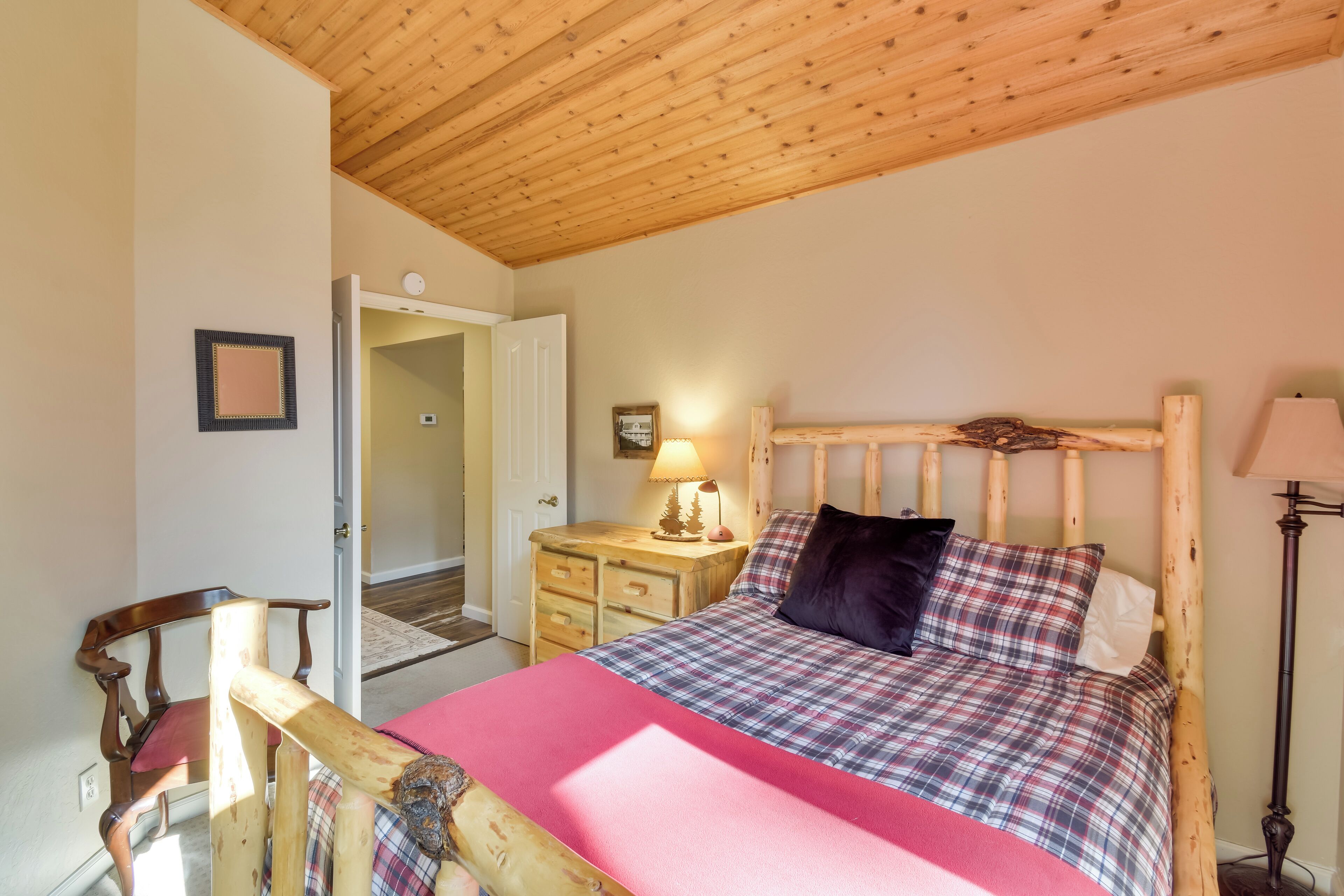 A bedroom with a wooden headboard, a bed with a plaid bedspread, a nightstand with a lamp, and a wooden chair.