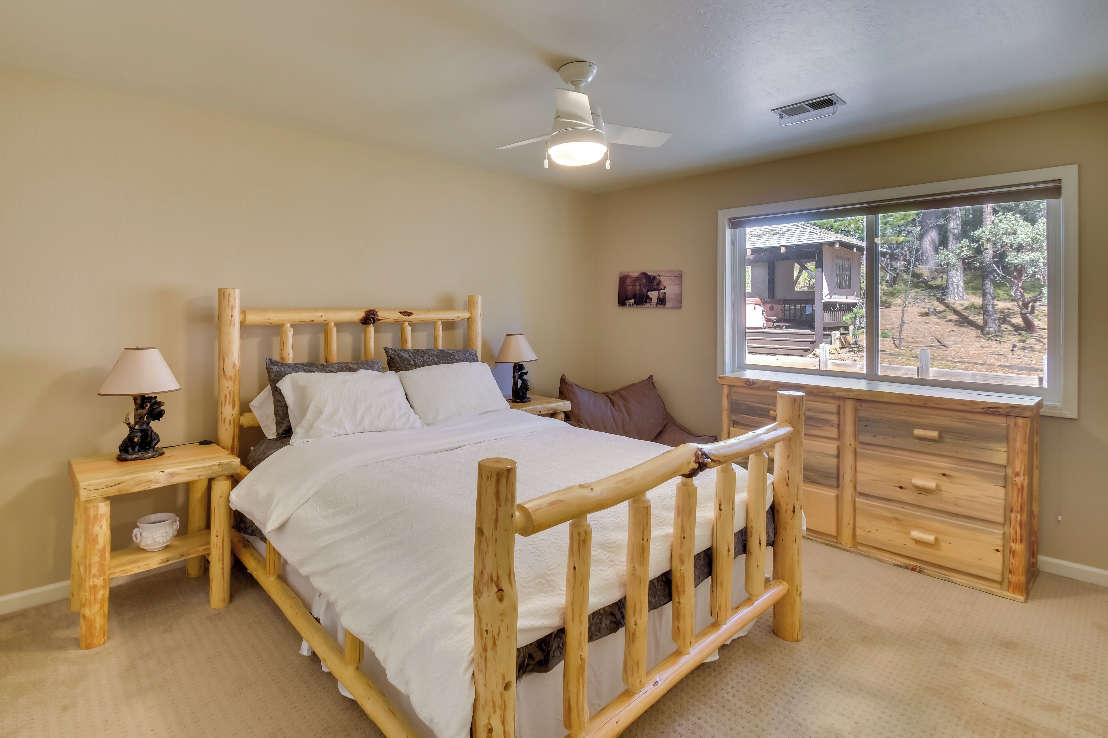 A bedroom with a wooden bed, two bedside tables with lamps, a window with a view of the outdoors, and a ceiling fan.