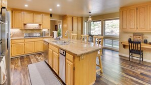 A spacious kitchen with wooden cabinets, a central island with a sink, a dishwasher, and a dining area with a table and chairs. - Hot Tub + Pool Table: Cabin Near Calaveras Park! (Arnold)