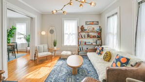 A spacious living room with a wooden floor, a round coffee table, a sofa with colorful cushions, a bookshelf, and a hanging light fixture. - Bright & Pet-friendly Home: 2 Mi to Harvard Square (Watertown)