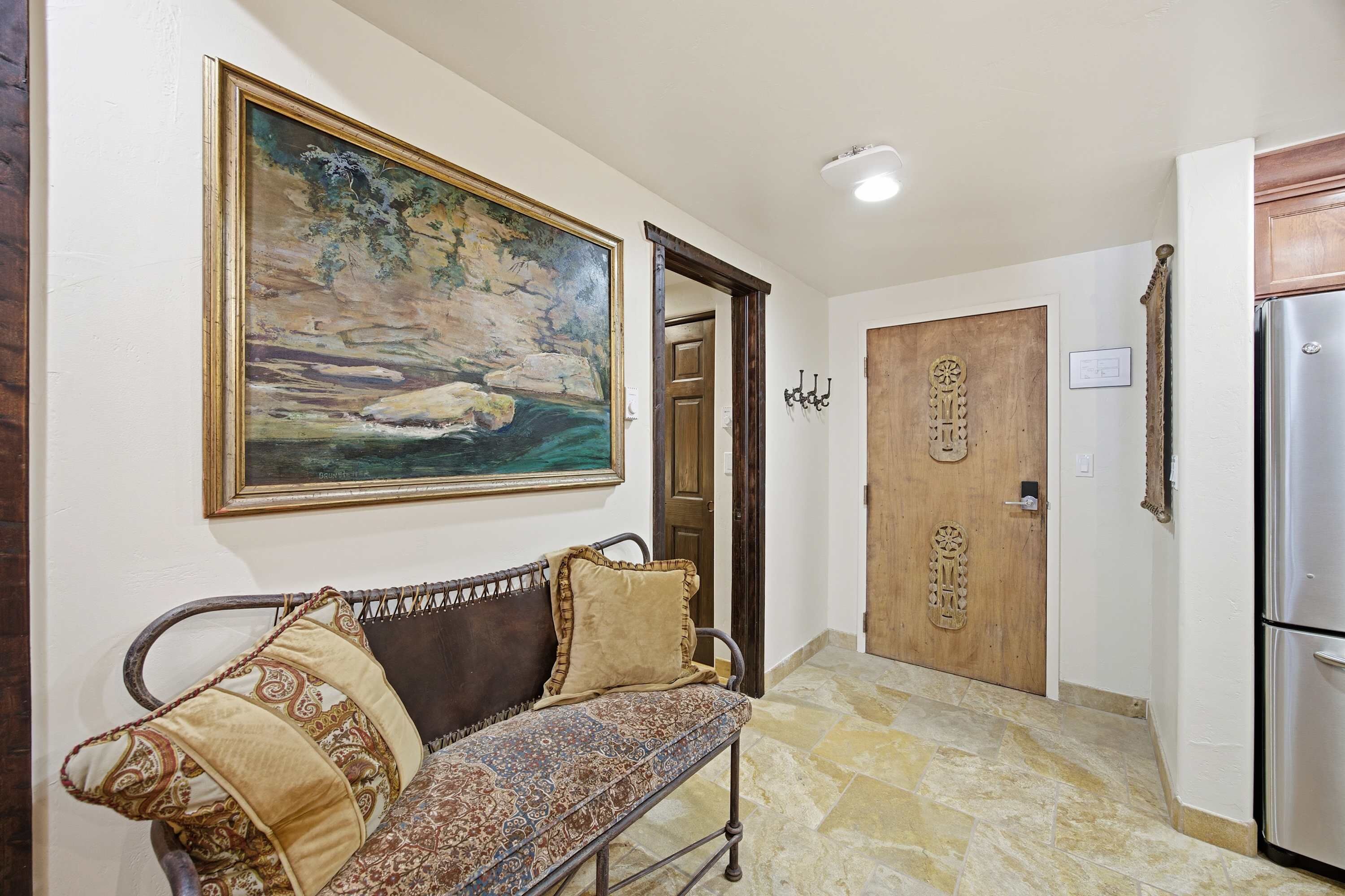 A hallway with a bench, a painting, a wooden door, and a refrigerator.