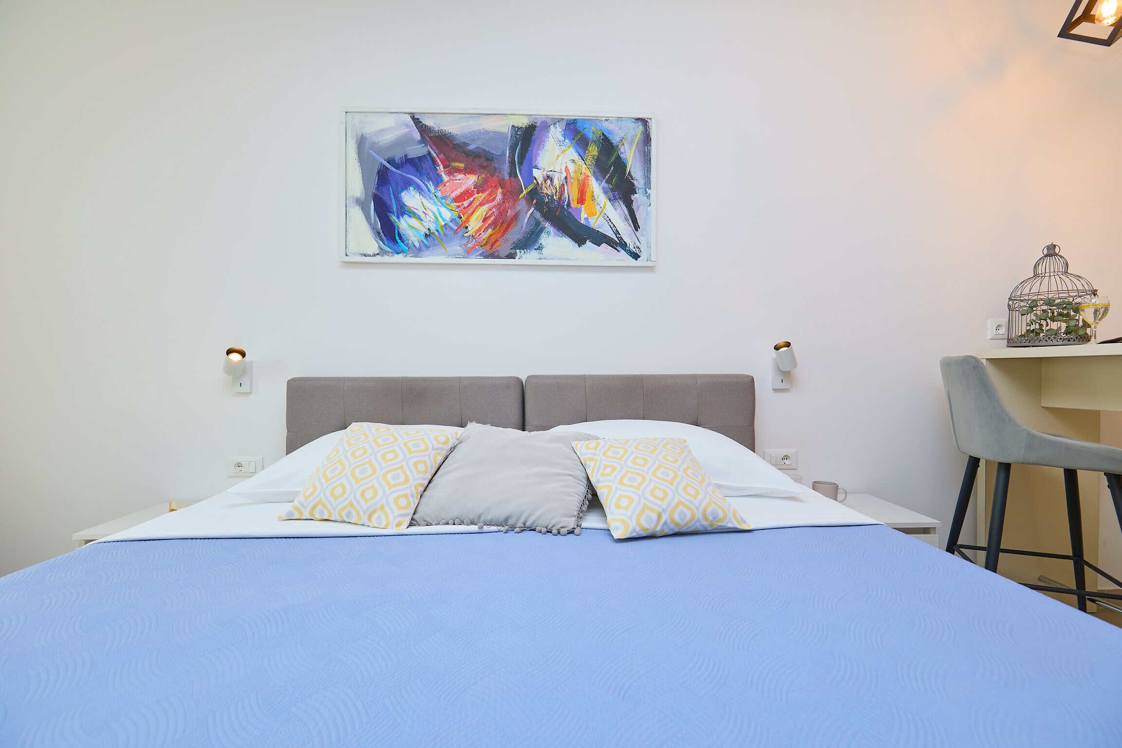 Ein ordentlich bezogenes Bett mit gemusterten Kissen, ein an der Wand hängendes abstraktes Gemälde und ein kleiner Tisch mit einer Dekoration.