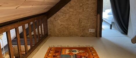 Une pièce avec un plafond en bois, un mur en pierre, un tapis coloré et une fenêtre avec des rideaux.
