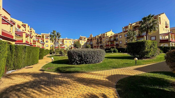 A courtyard with a brick pathway, neatly trimmed hedges, and a row of yellow buildings with balconies. - GUA001 (Ayamonte)
