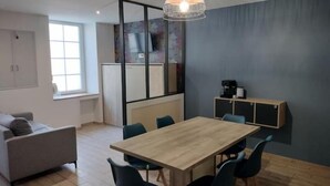 A modern kitchen with a wooden table, blue chairs, a hanging light fixture, and a window. - Logement 3 (Les Epesses)