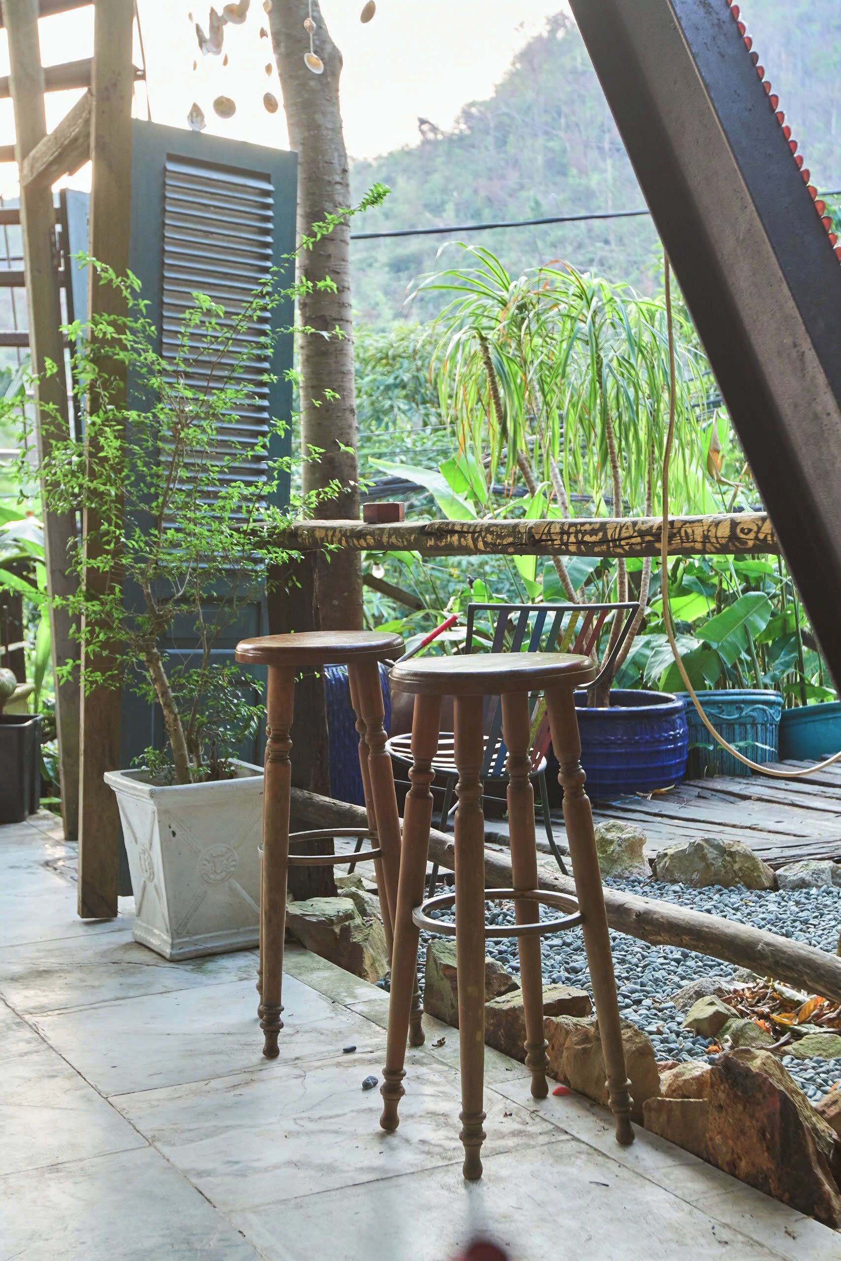 A wooden chair and table set on a tiled patio with potted plants and a view of a lush garden.