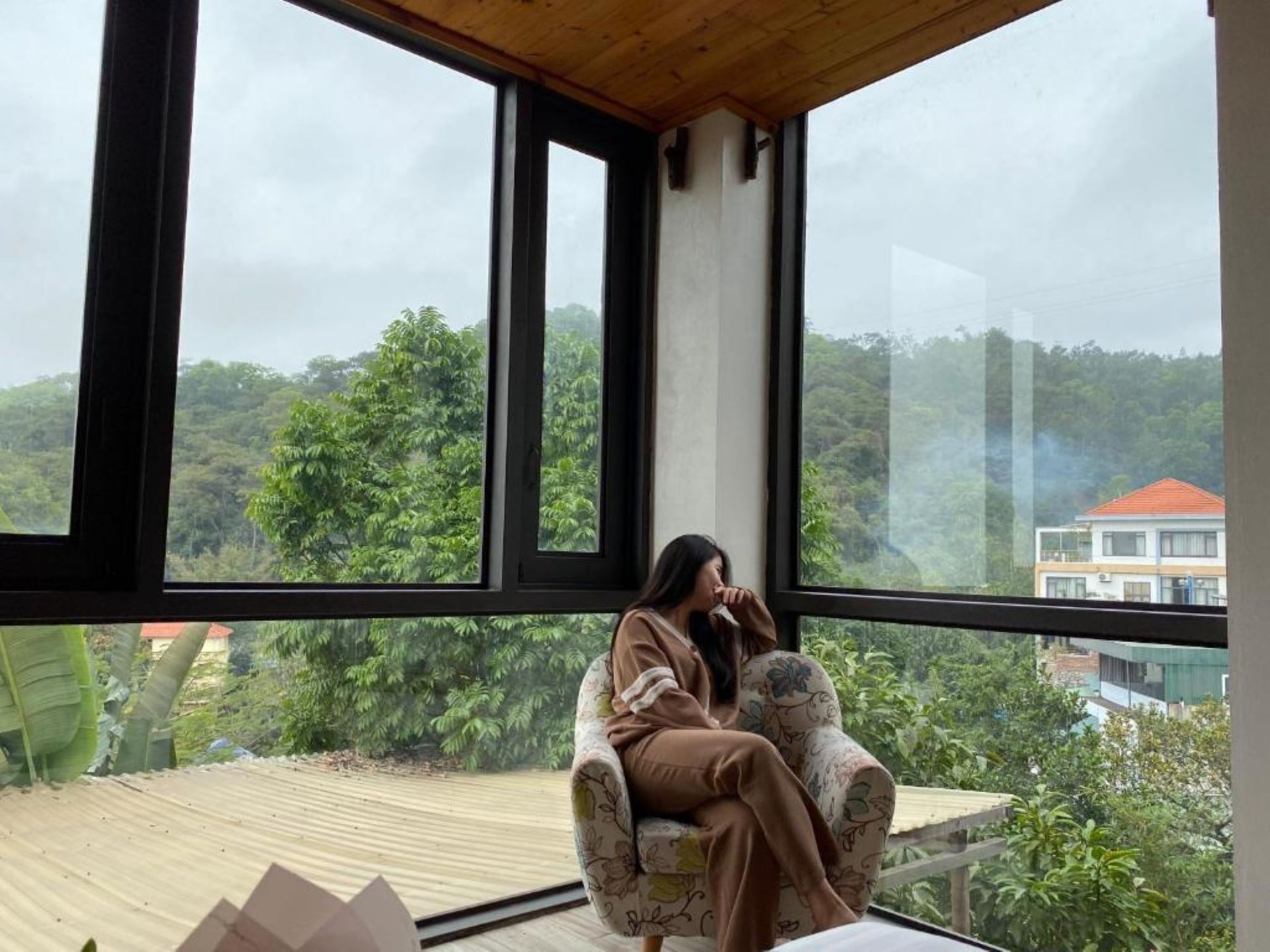 Ein Zimmer mit großen Fenstern und Blick auf üppiges Grün sowie ein weiter Blick auf ein Gebäude in der Ferne.