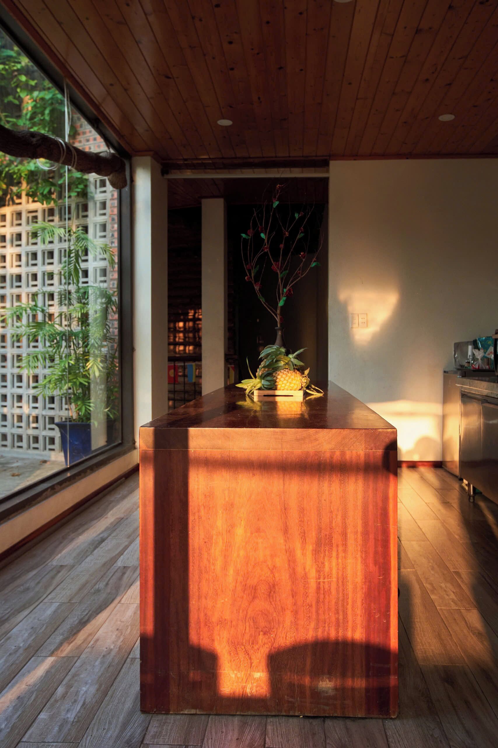Un mostrador de recepción de madera con una planta en maceta, vista a un jardín de bambú a través de una puerta de vidrio y una cocina moderna al fondo.
