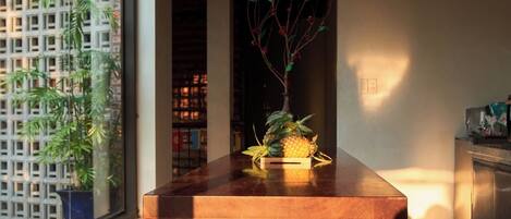 Un mostrador de recepción de madera con una planta en maceta, vista a un jardín de bambú a través de una puerta de vidrio y una cocina moderna al fondo.