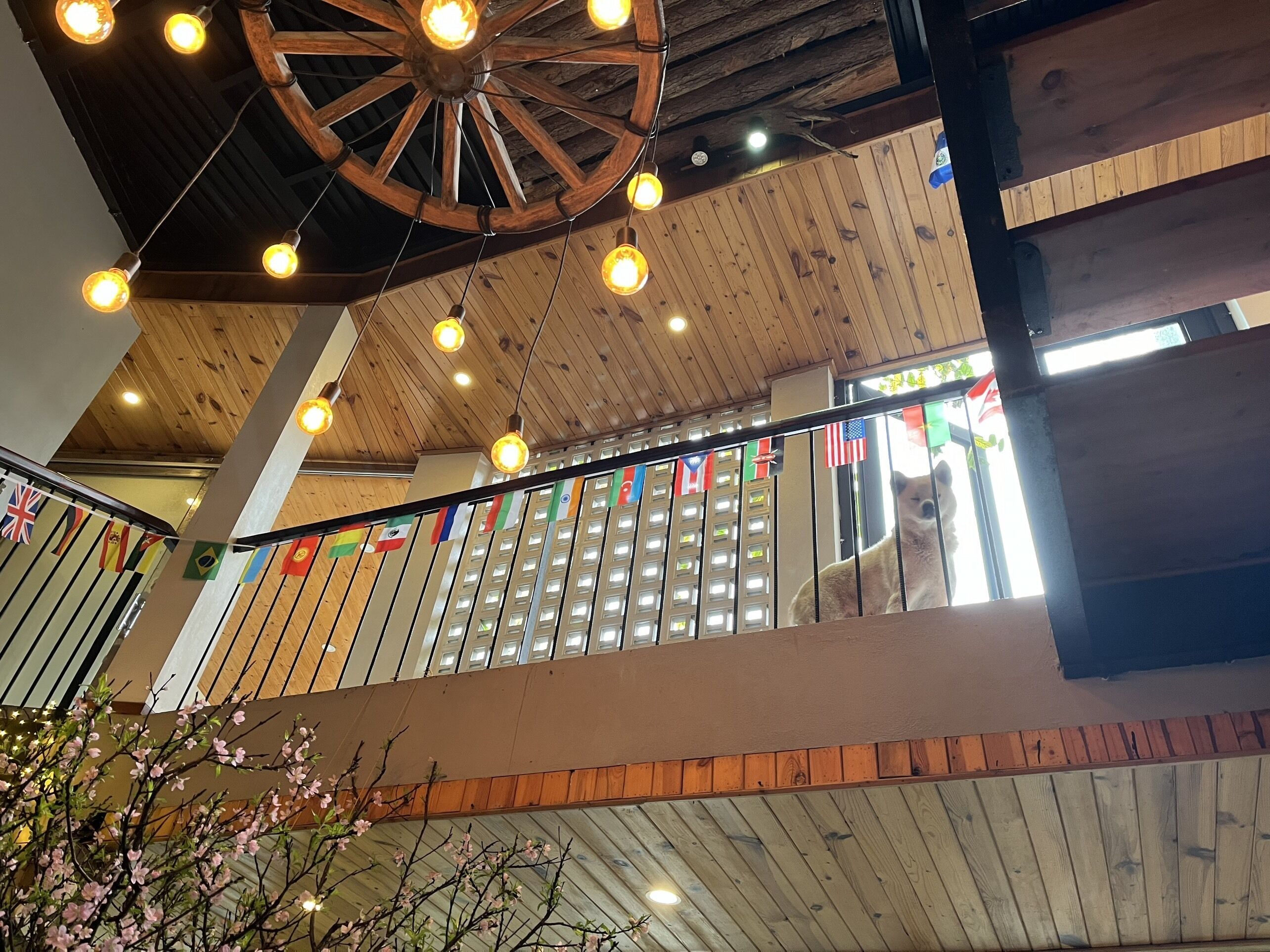 A multi-level wooden interior with a central wooden wheel structure, multiple light fixtures, and a balcony railing adorned with various national flags.
