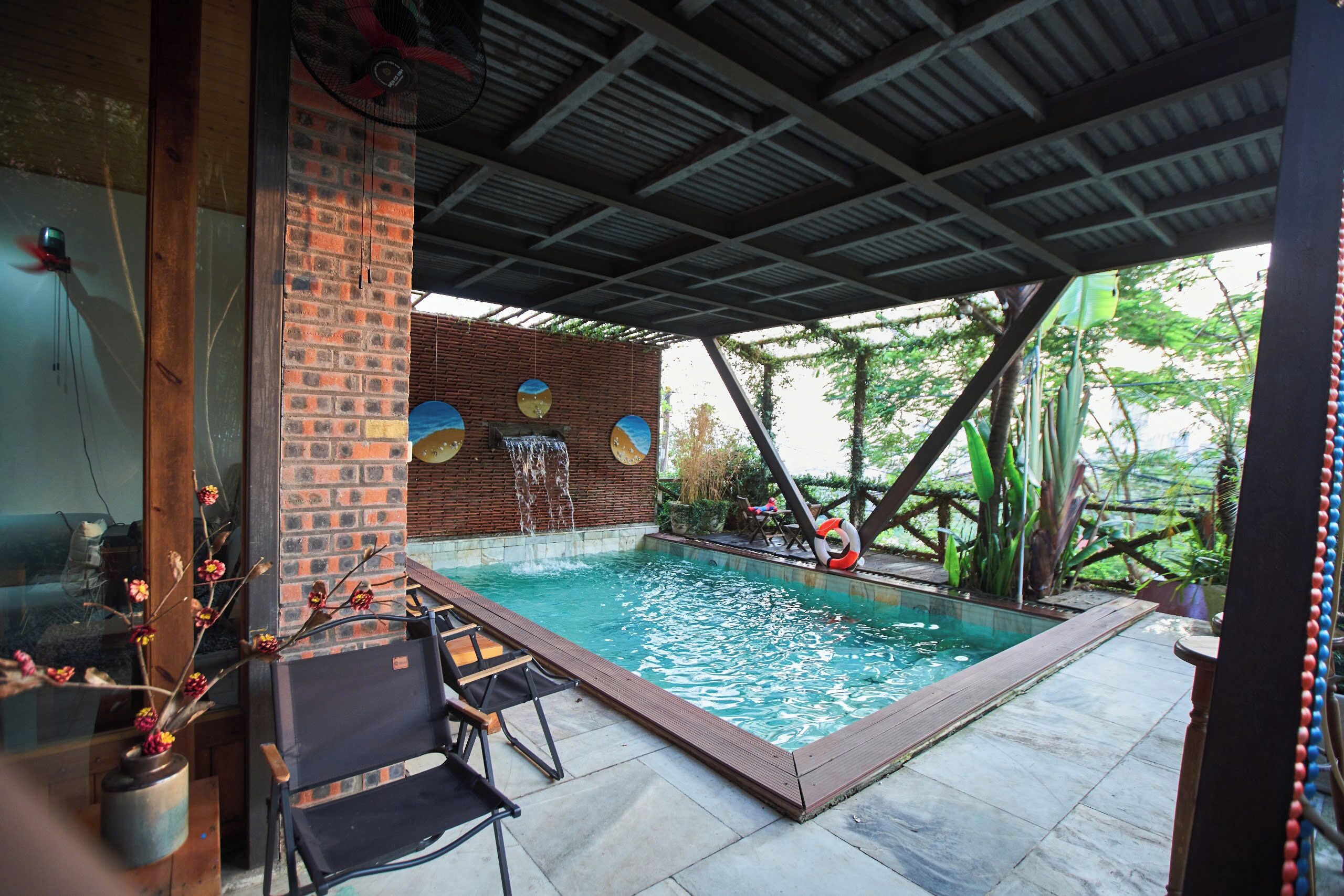 Área de piscina com muro de tijolos, deck de madeira e espaço coberto com cadeiras.
