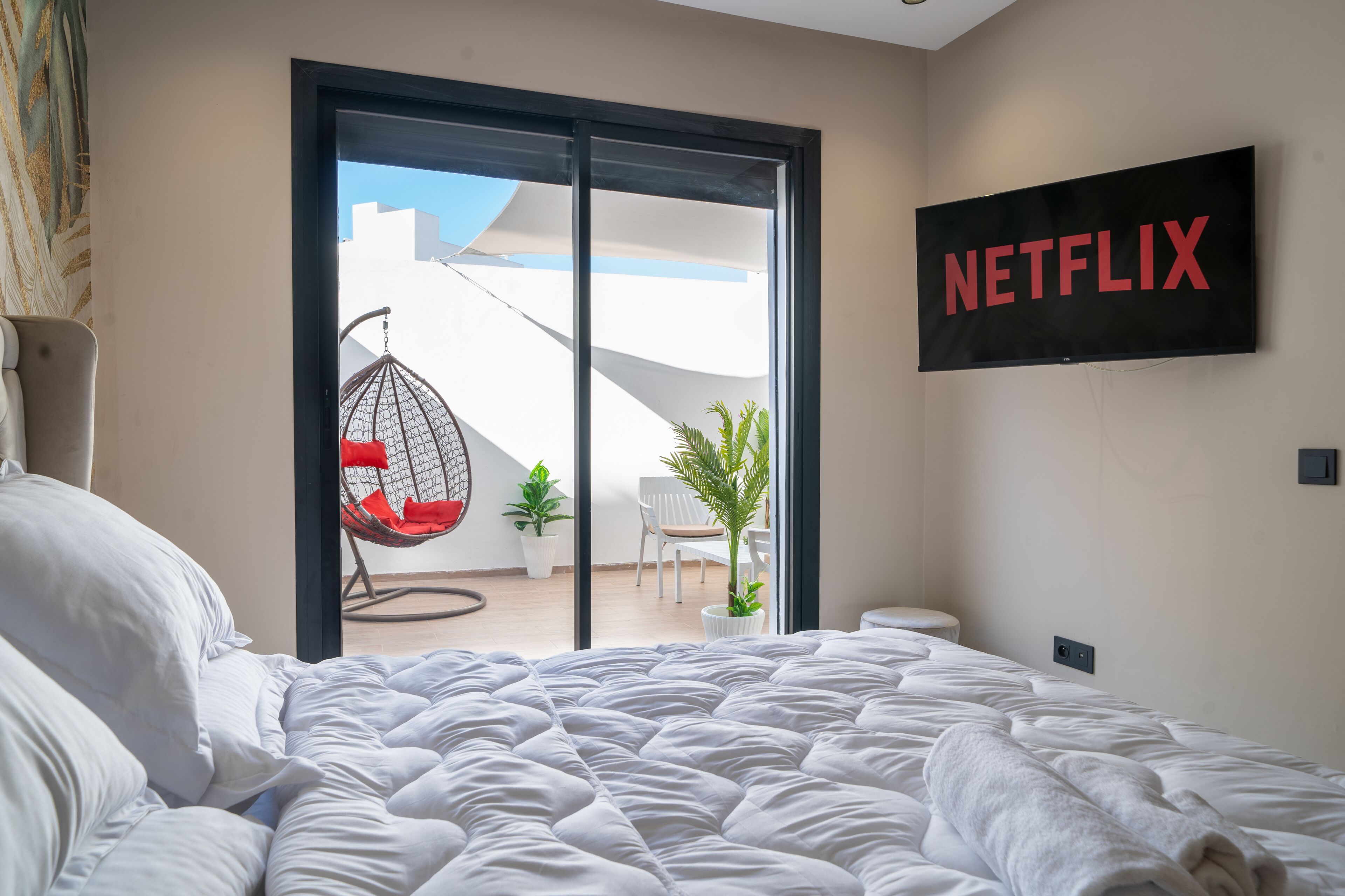 Et soveværelse med en stor seng, en skydedør til en balkon, en hængekøjsstol og et vægmonteret fjernsyn, der viser Netflix logoet.