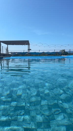 Una piscina trasparente con fondo piastrellato, una zona coperta con tetto e una recinzione sullo sfondo.