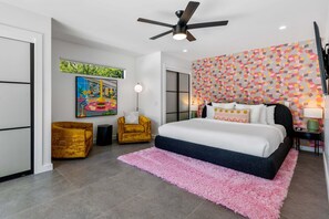 Une chambre à coucher avec un lit, un ventilateur de plafond, un tapis rose, un fauteuil jaune et un mur aux couleurs vives.