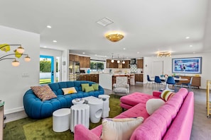 Un salon moderne avec un canapé d’angle bleu, un canapé rose et un fauteuil à carreaux. On y trouve également un coin repas avec une table et des chaises, et une cuisine en arrière-plan.