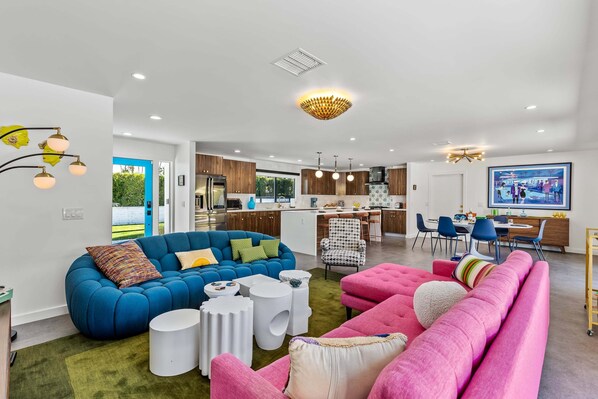 Una sala de estar moderna con un sofá seccional azul, un sofá rosa y un sillón a cuadros. Hay un comedor con mesa y sillas, y una cocina al fondo.