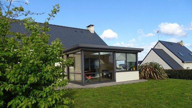 Une maison moderne dotée d’une véranda en verre, d’une pelouse bien entretenue et d’une maison voisine avec un toit en pente.
