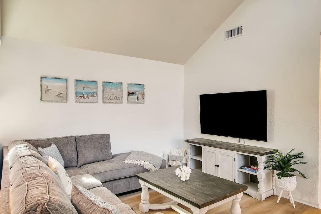 A living room with a grey sofa, a coffee table, a flat-screen TV, and wall art depicting beach scenes.
