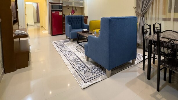 Una sala moderna con un sofá azul, un sillón amarillo, una mesa de comedor con sillas, un refrigerador rojo y una alfombra estampada.