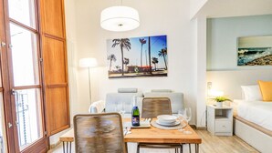 A dining area with a wooden table, two wicker chairs, a painting of palm trees, a bed with a white and yellow pillow, and a nightstand with a lamp. - The Green Oasis by Hello Homes Sitges (Sitges)