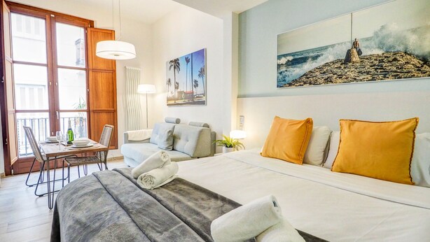 A hotel room with a bed, a dining table with chairs, a sofa, and a painting of a beach scene. - The Green Oasis by Hello Homes Sitges (Sitges)