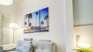 A modern hotel room with a grey sofa, a wall-mounted painting of palm trees, a floor lamp, and a potted plant. - The Green Oasis by Hello Homes Sitges (Sitges)