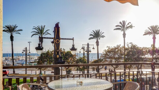 A balcony with a round glass table and wicker chairs, overlooking a marina with boats and palm trees. - Soleil by Hello Homes Sitges (Sitges)