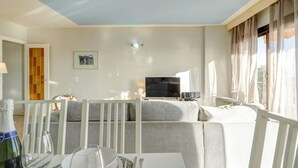A modern living room with a grey sofa, a flat-screen TV, and a dining area with a table set for two. - Soleil by Hello Homes Sitges (Sitges)