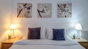 A neatly made bed with white and dark pillows, flanked by bedside tables with lamps and small potted plants. Three floral artwork pieces adorn the wall above the bed. - Soleil by Hello Homes Sitges (Sitges)