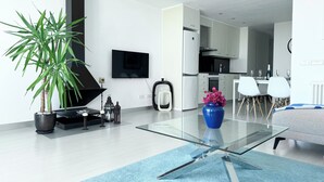 A modern living room with a glass coffee table, a large potted plant, a blue vase with flowers, and a flat-screen TV mounted on the wall. - Vistas Del Mar by Hello Homes Sitges (Sitges)