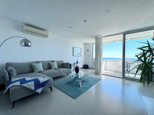 A modern living room with a grey sofa, a glass coffee table, and a view of the sea through large windows.