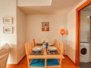 Ein Essbereich mit einem Holztisch, gedeckt für vier Personen, umgeben von Stühlen. Darüber hängt ein Wandbild, rechts steht eine Stehlampe und im Hintergrund ist eine Waschmaschine zu sehen.