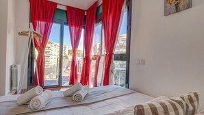 Habitación de hotel con cama, cortinas rojas, ventana con vista a edificios y un cuadro en la pared.